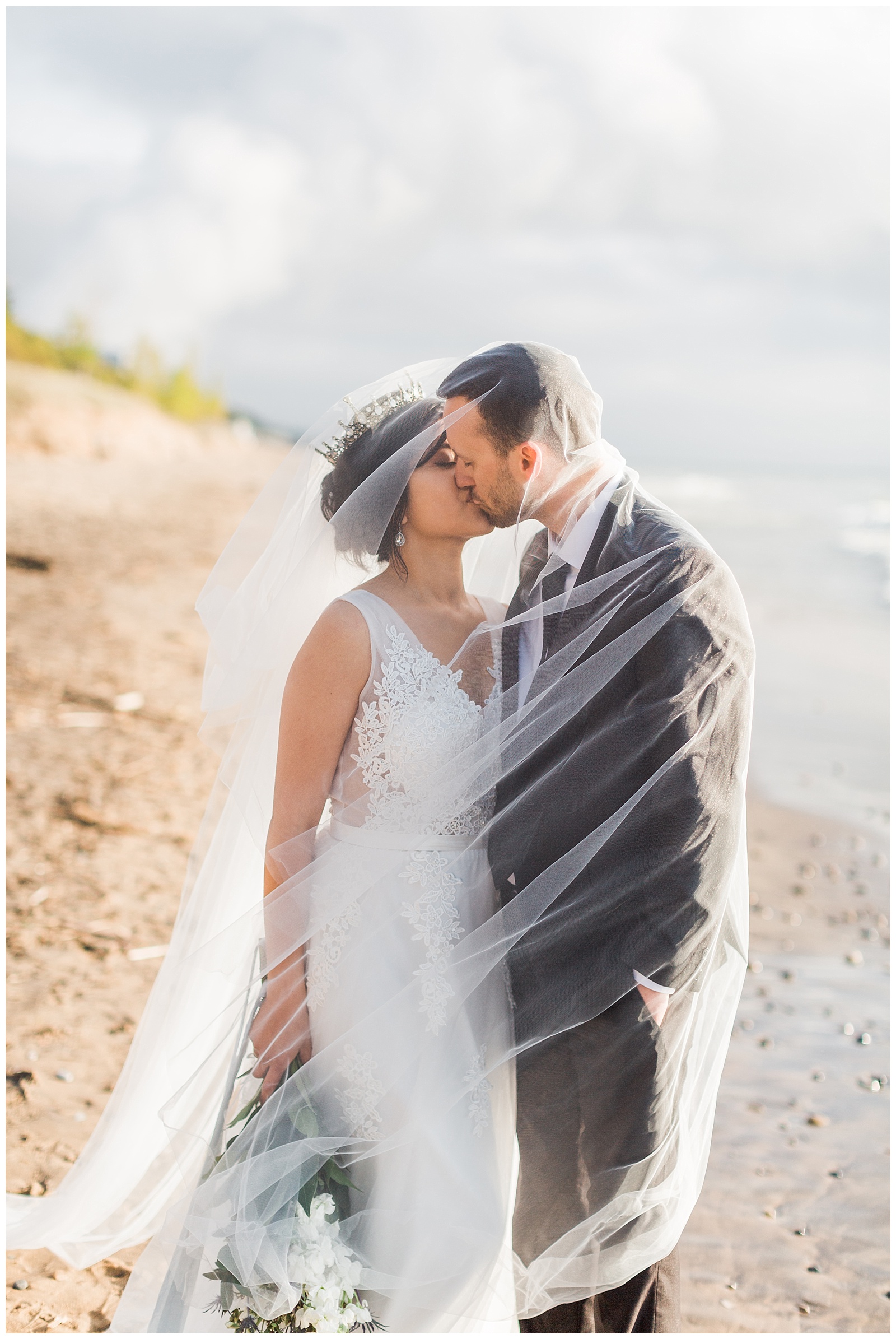 Bride and groom kiss under veil during Northern Michigan beach wedding.