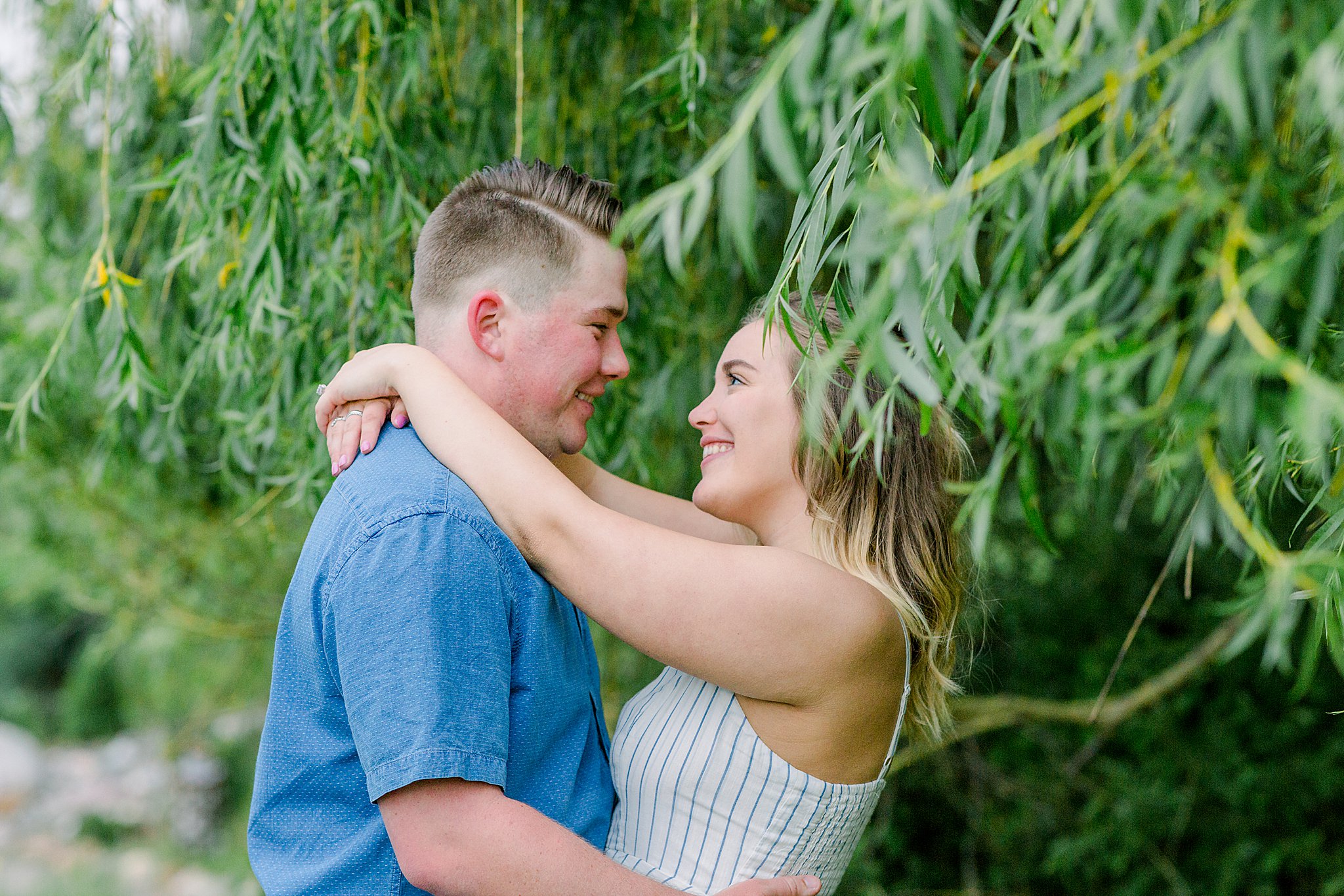 Couple smiles at each other under a willow tree during Grand Traverse Bay engagement session.