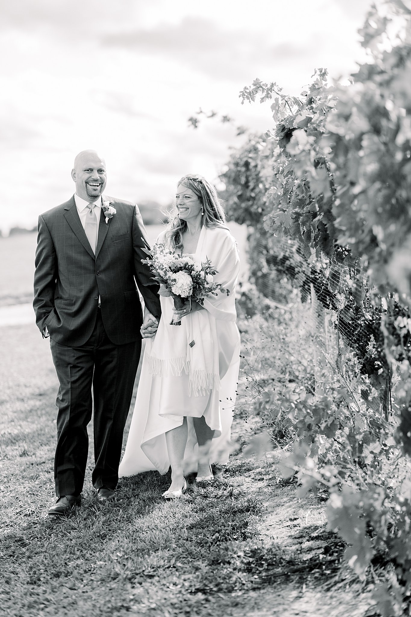 Black and white image of bride and groom laughing while walking through vineyards during their Aurora Cellars wedding.