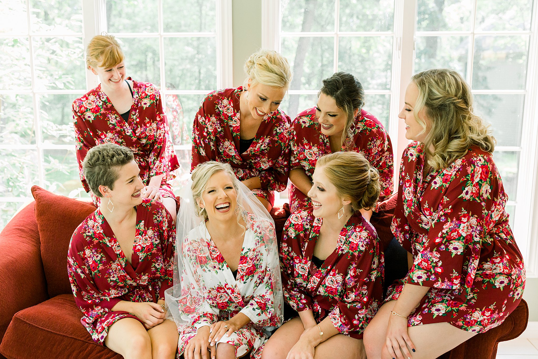 Bridesmaids surround bride on red couch laughing together while getting ready for Post Family Farm Wedding