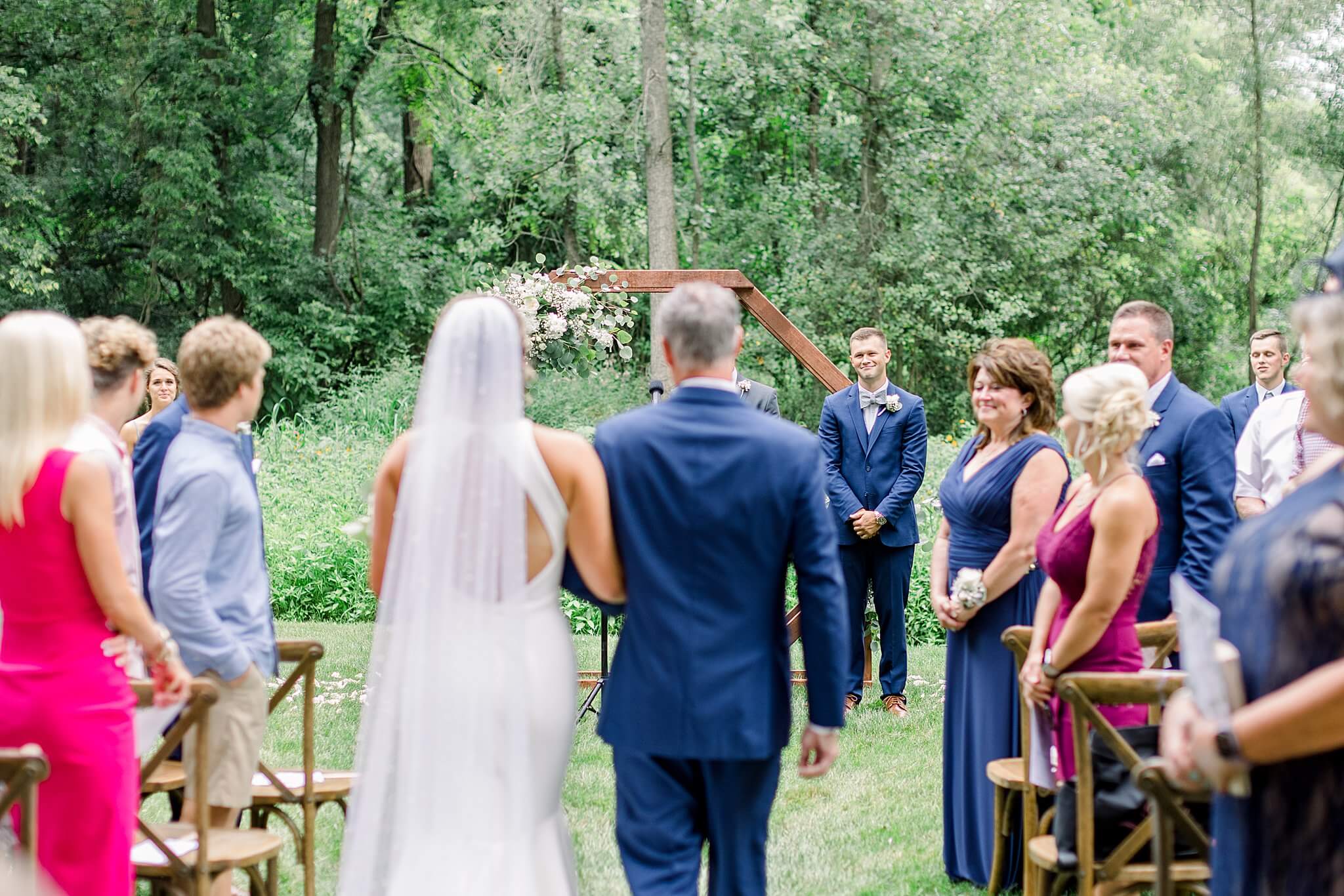 Groom smiles as his bride walks down the aisle with her dad during their summer backyard wedding in Northern Michigan.