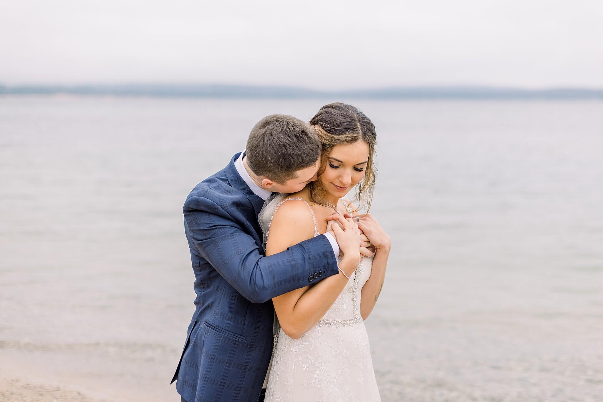 Groom kisses bride's neck during pictures on the beach for their Intimate Northern Michigan Wedding day. 