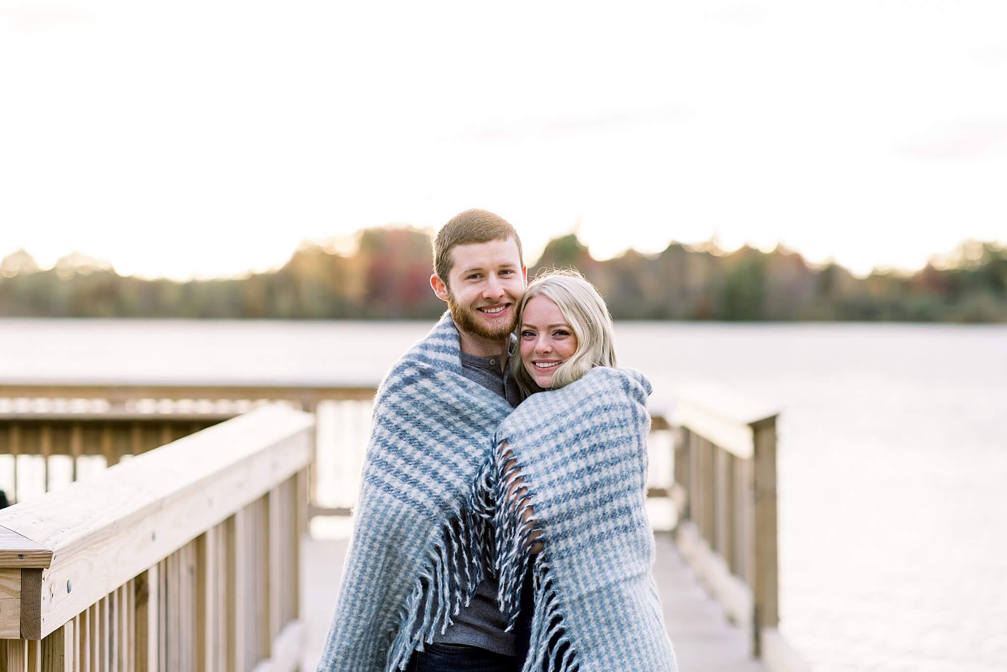 Bride and groom snuggle under cozy blanket on a dock during M22 Fall Engagement Session in Northern Michigan.