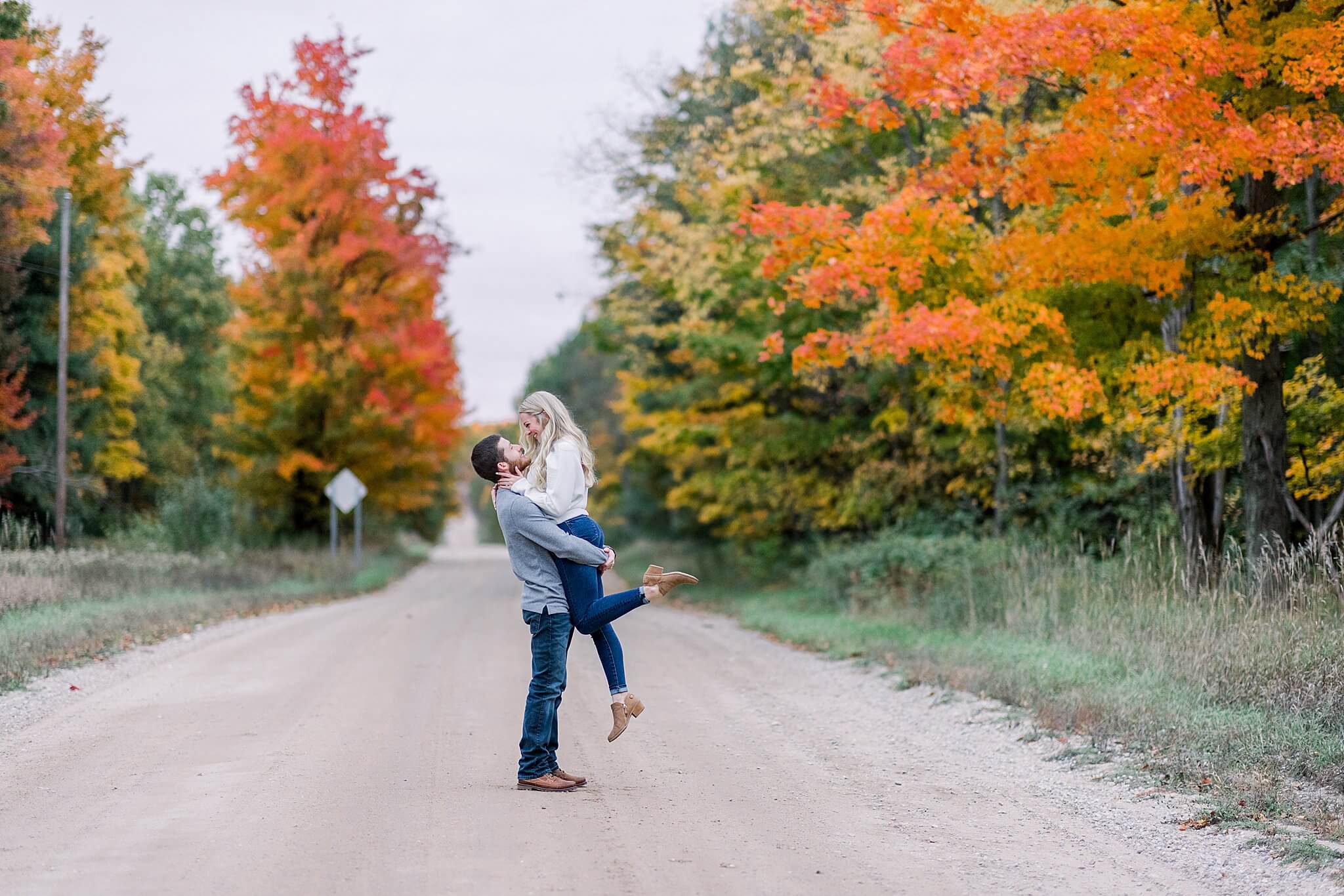 Groom lifts bride along colorful fall road during M22 Fall Engagement Session in Northern Michigan.