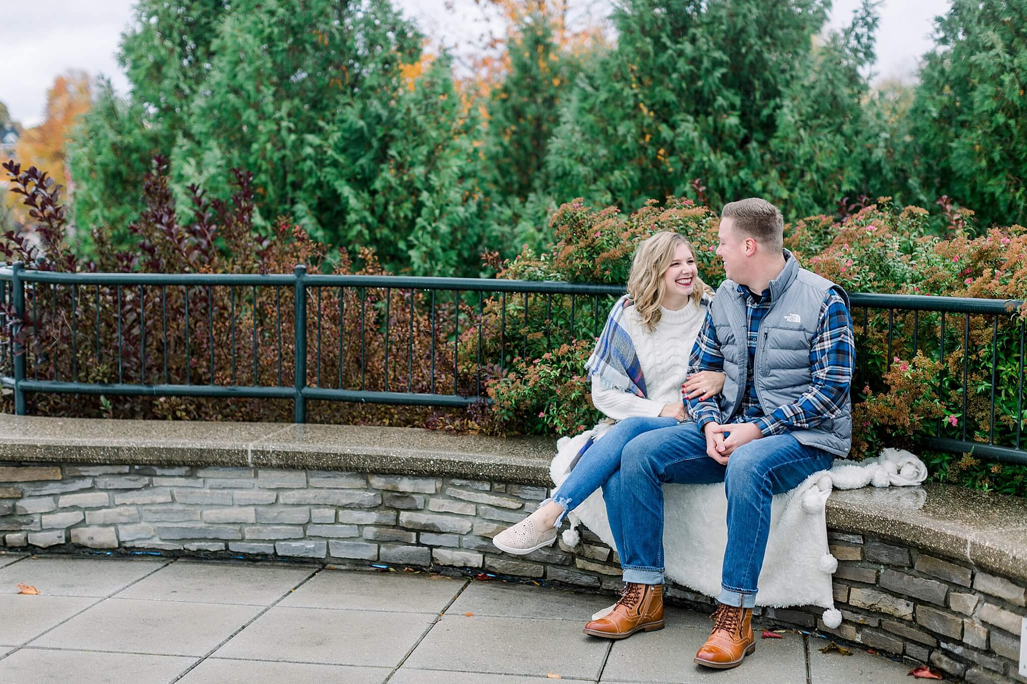 Bride and groom snuggle together on stone wall during rainy fall engagement session in Charlevoix, Michigan.