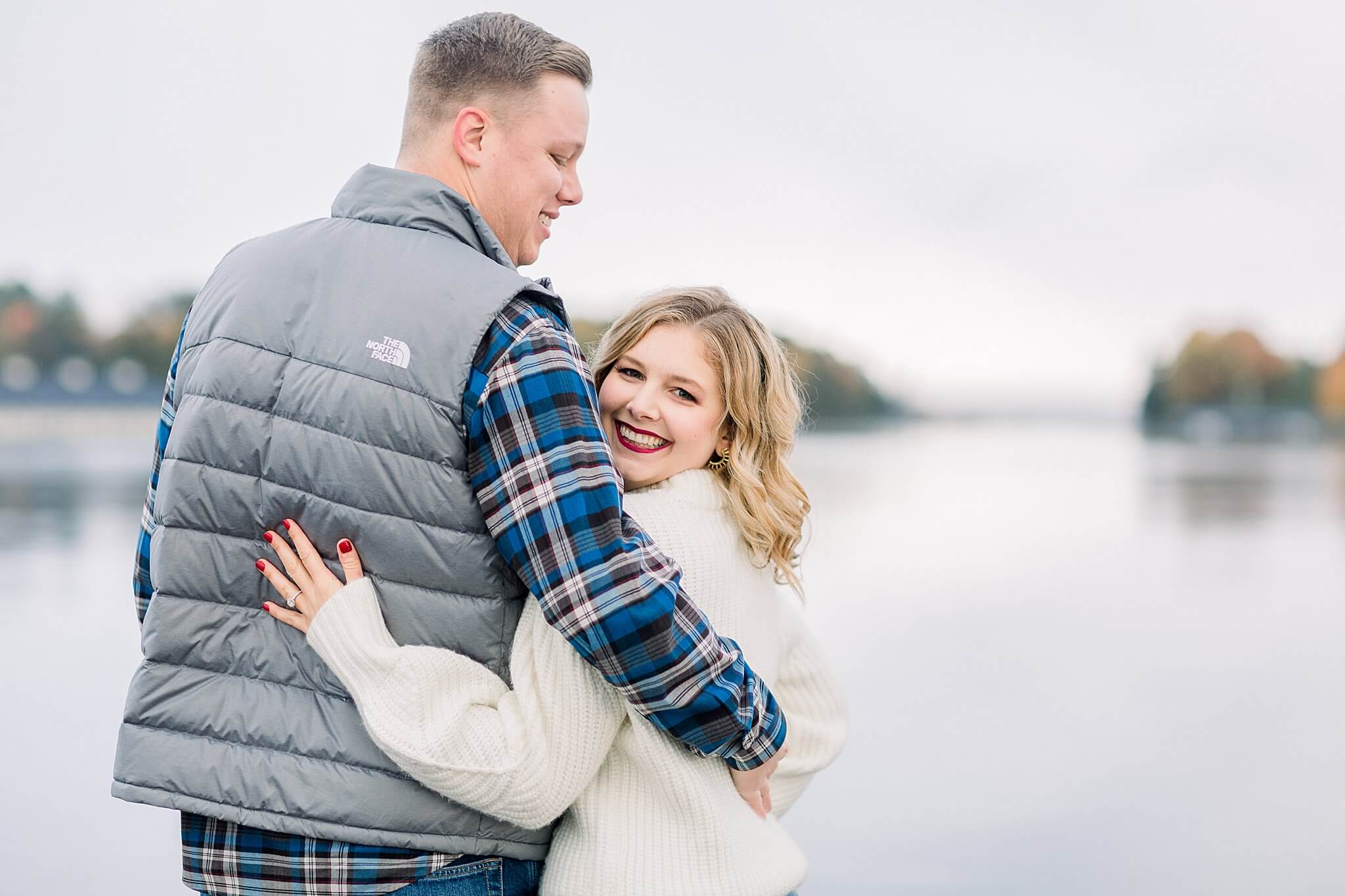 Groom smiles at bride on Lake Charlevoix during rainy fall engagement session in Charlevoix, Michigan.