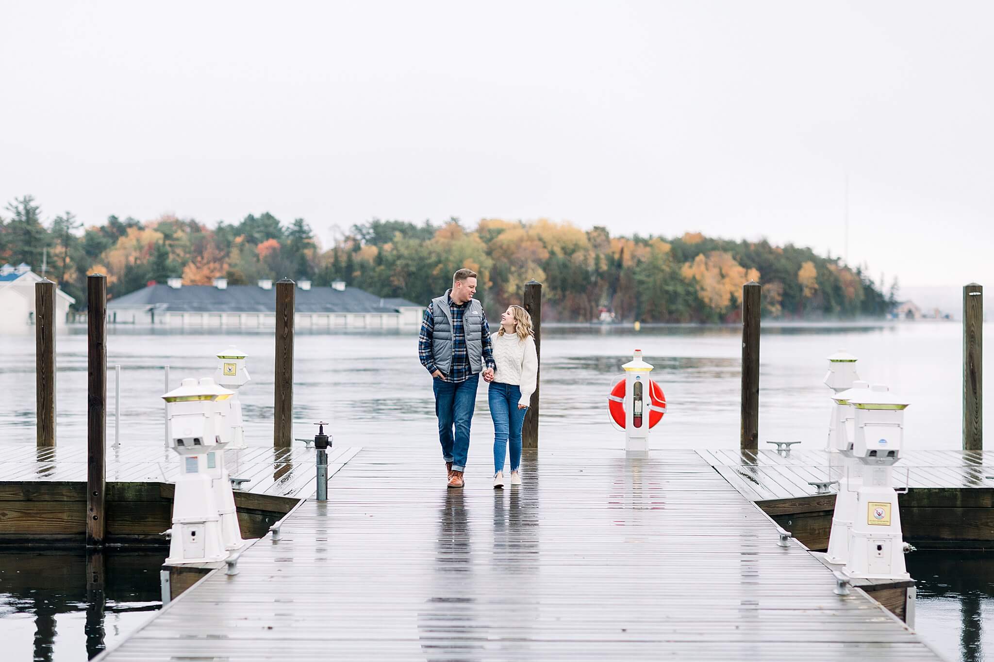 Bride and groom walk together on rainy dock during rainy fall engagement session in Charlevoix, Michigan.