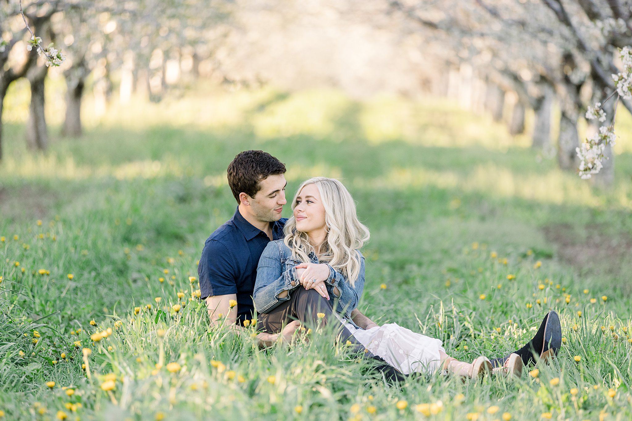Couple sits in a field of wildflowers in a cherry orchard during their cherry blossom engagement session in Traverse City, Northern Michigan. 