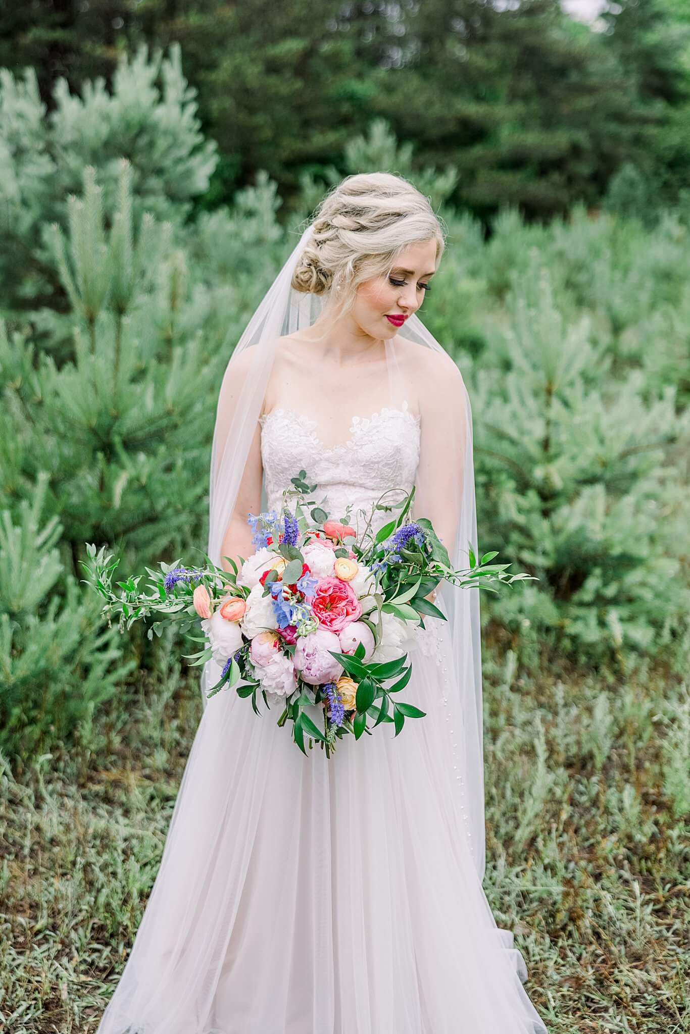 Sweet, romantic bride poses in front of evergreen trees with a bright, colorful bouquet on her Traverse City wedding daya.