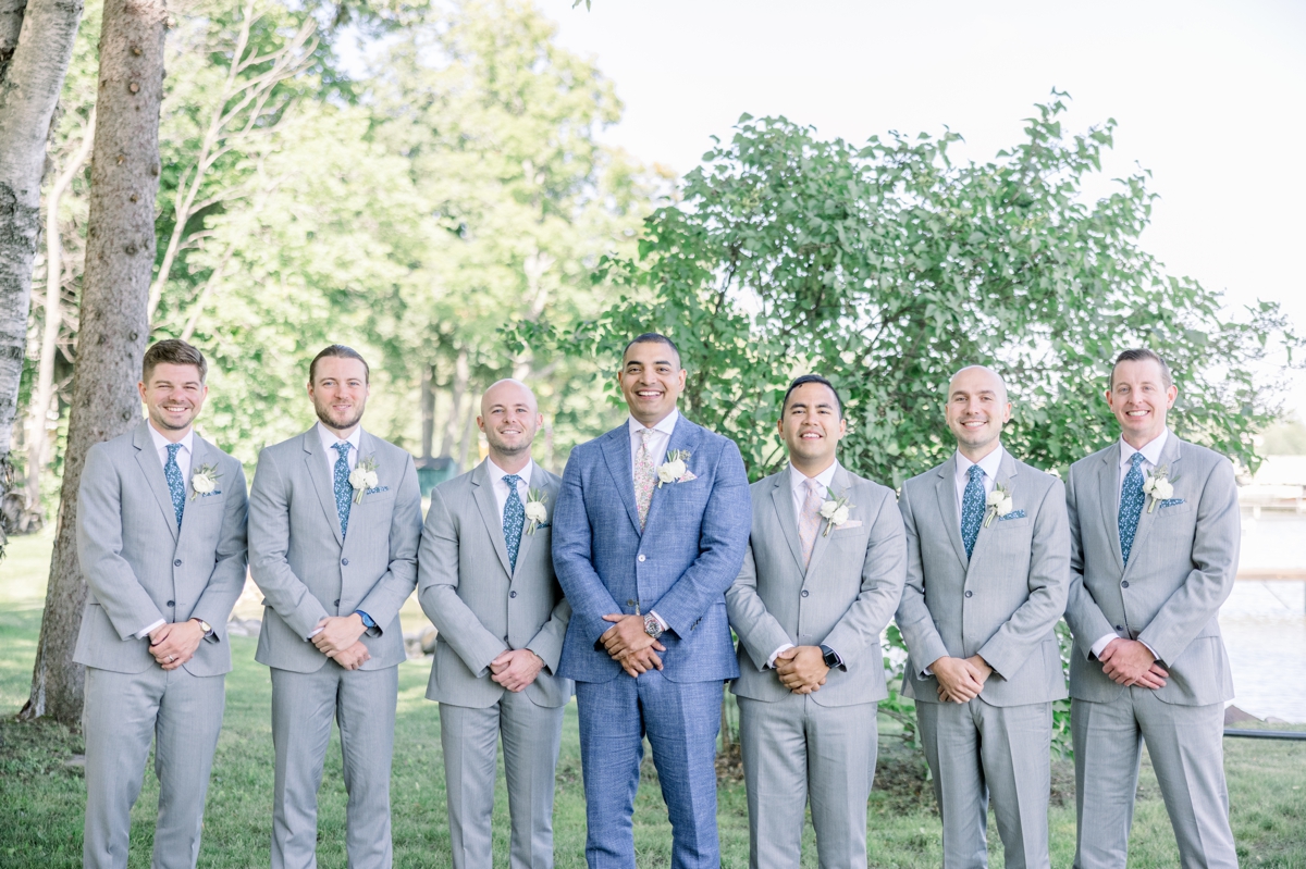 Andres smiling in his blue suit as he stands with his groomsmen in their grey suits on the lawn of his lake house on this wedding day.