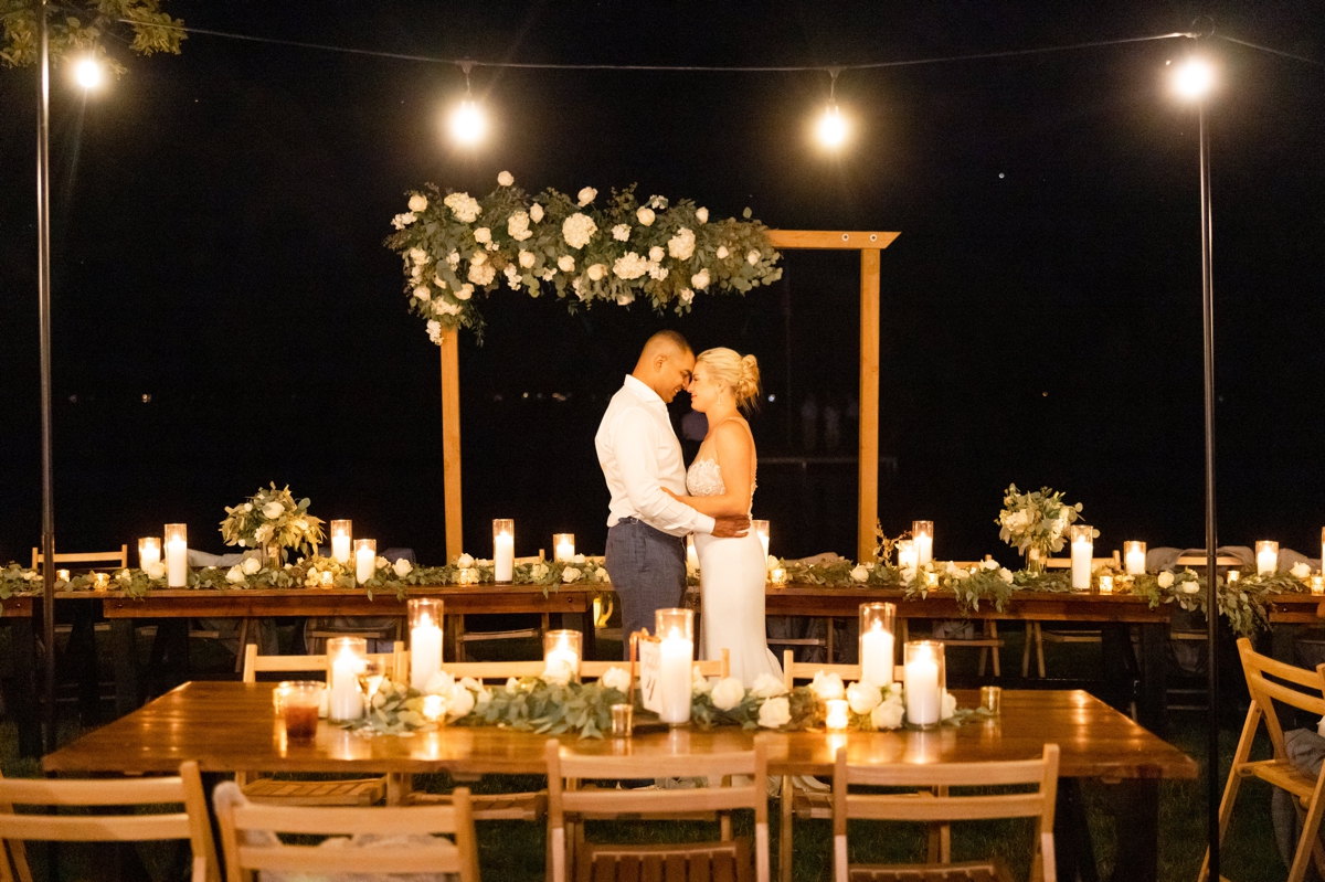 Caitlin and Andres standing in front of their head table arch with their foreheads together taking in the last moments of their Northern Michigan lake house wedding as candles glow on the tables around them.