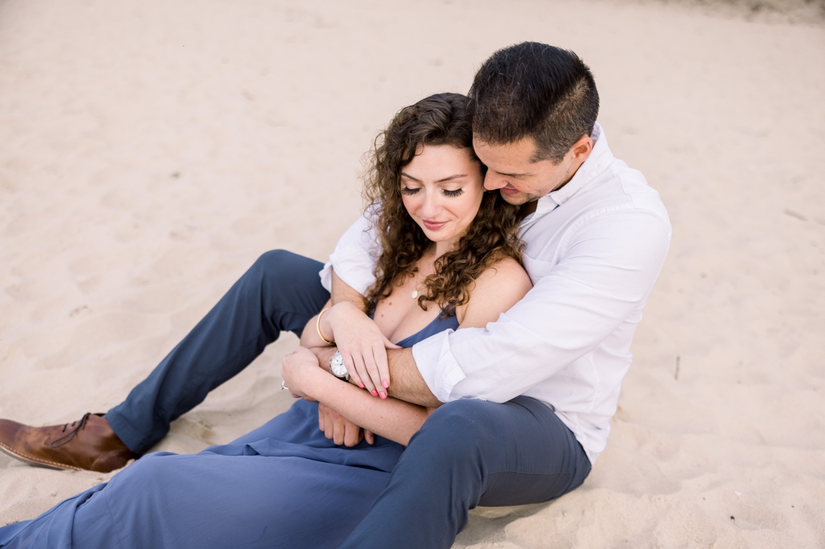 Alex hugging Marissa from behind while they sit in the sand during their engagement session.