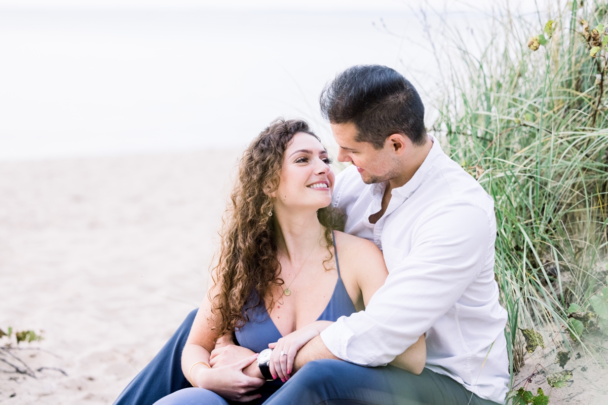 Marissa and Alex smiling at each other while they sit in the sand during their beach engagement session with Mandie Forbes Photography.