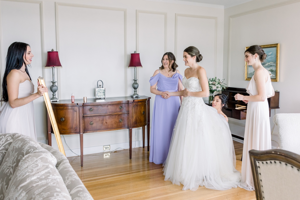 The bride standing with her sisters and mom while she looks at herself in a mirror in her wedding dress.