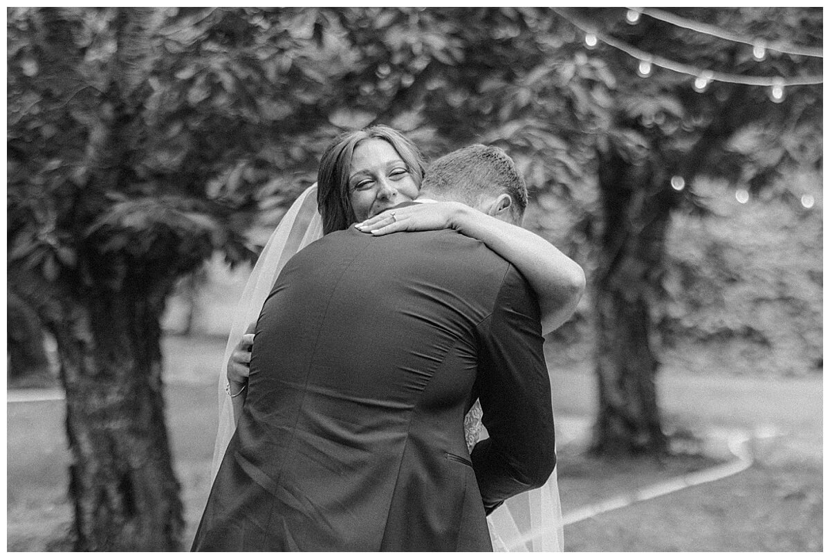 Bride hugs groom in emotional black and white photograph. They are surrounded by cherry trees in an orchard located at Bay View by Gallagher Farms in Traverse City, Northern Michigan.