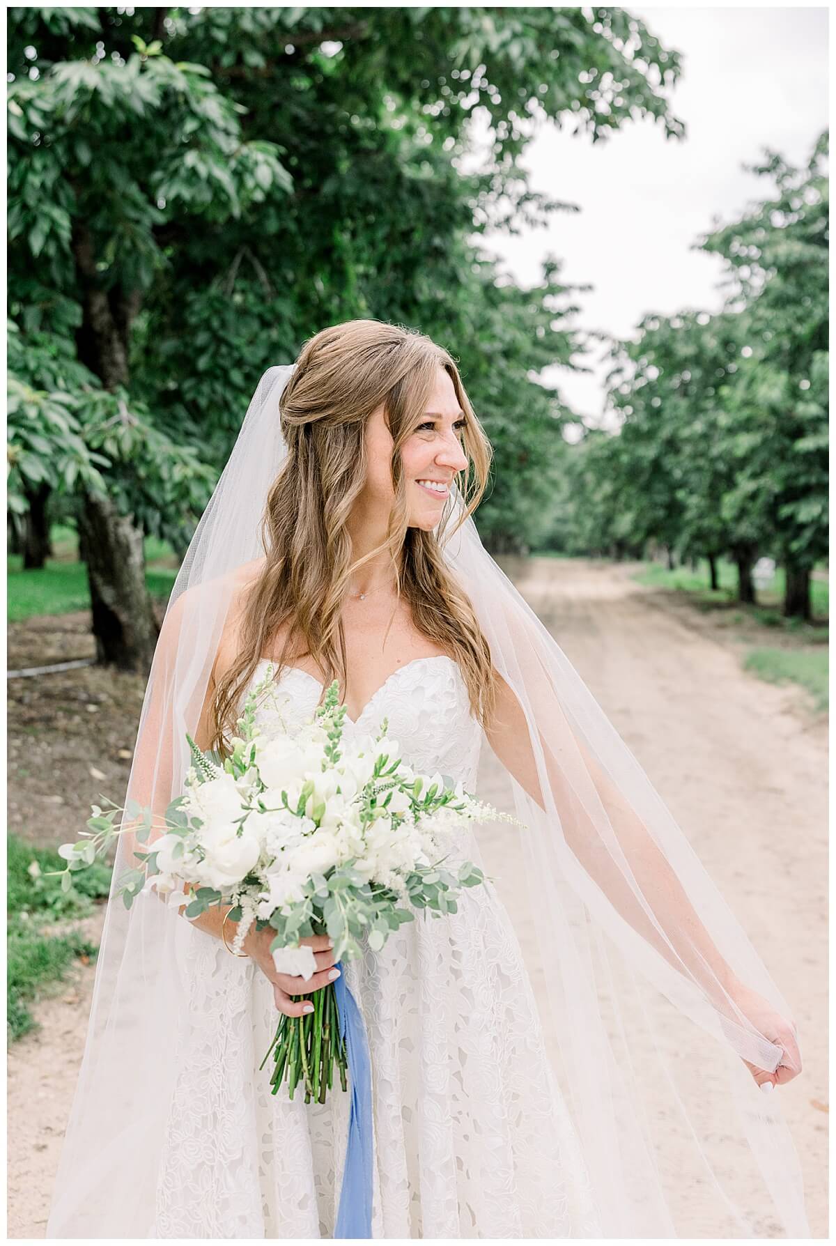 Beautiful bridal portrait of a bride wearing a stunning veil and a custom lace dress while holding a classic white bridal bouquet in the cherry orchards at Bay View in Traverse City.