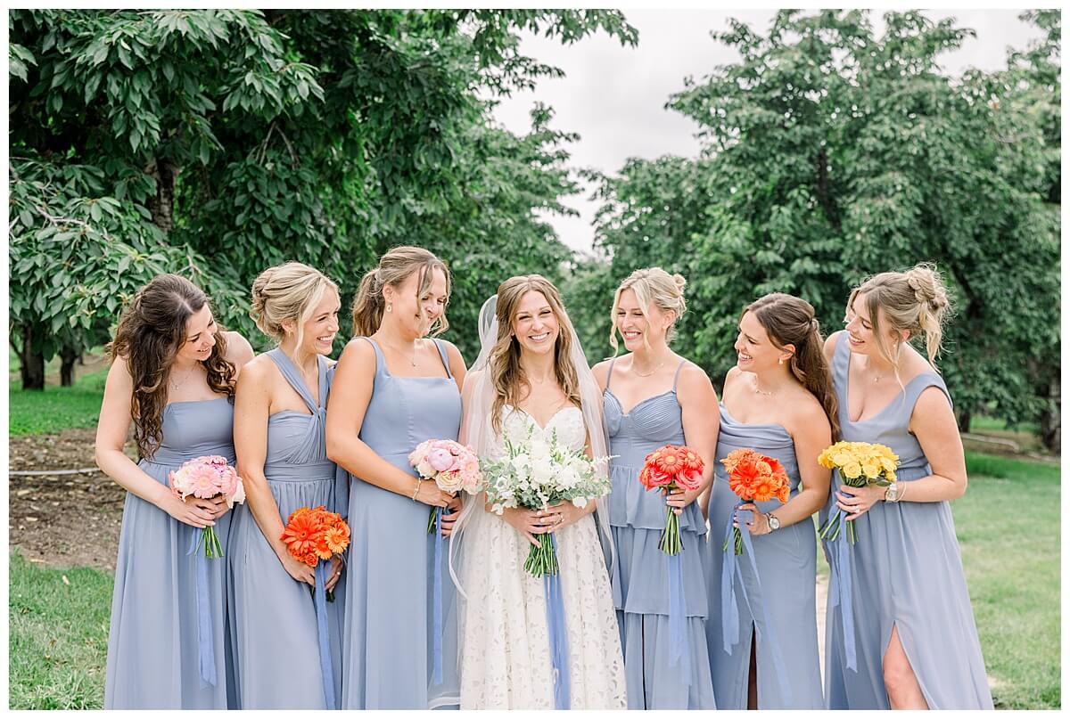 A stunning bride with her bridesmaids who are all weary dusty blue dresses all smiling at each other in the cherry orchards on Leelanau Peninula before her emotional Bay View wedding in Traverse City.