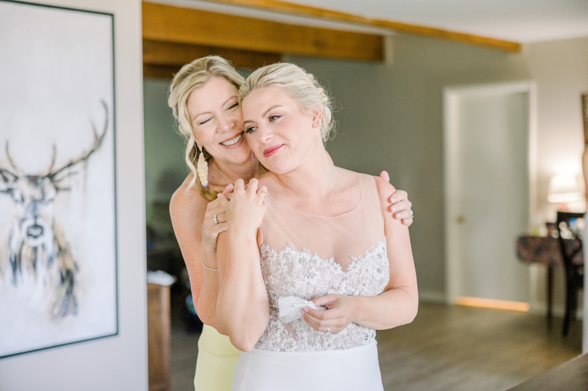 Caitlin being hugged from behind by her mom as they get her ready for her Northern Michigan Lake House wedding.