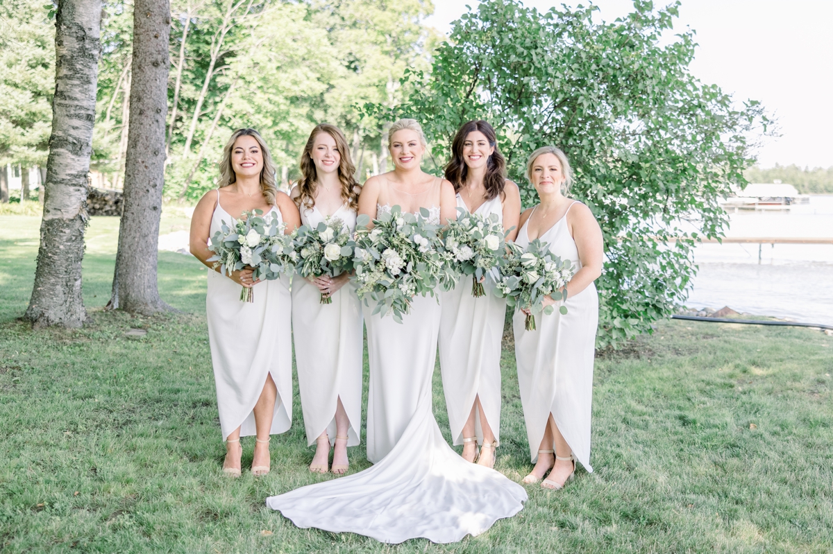 Caitlin smiling with her bridesmaids in light cream dresses at the lake house on her wedding day.