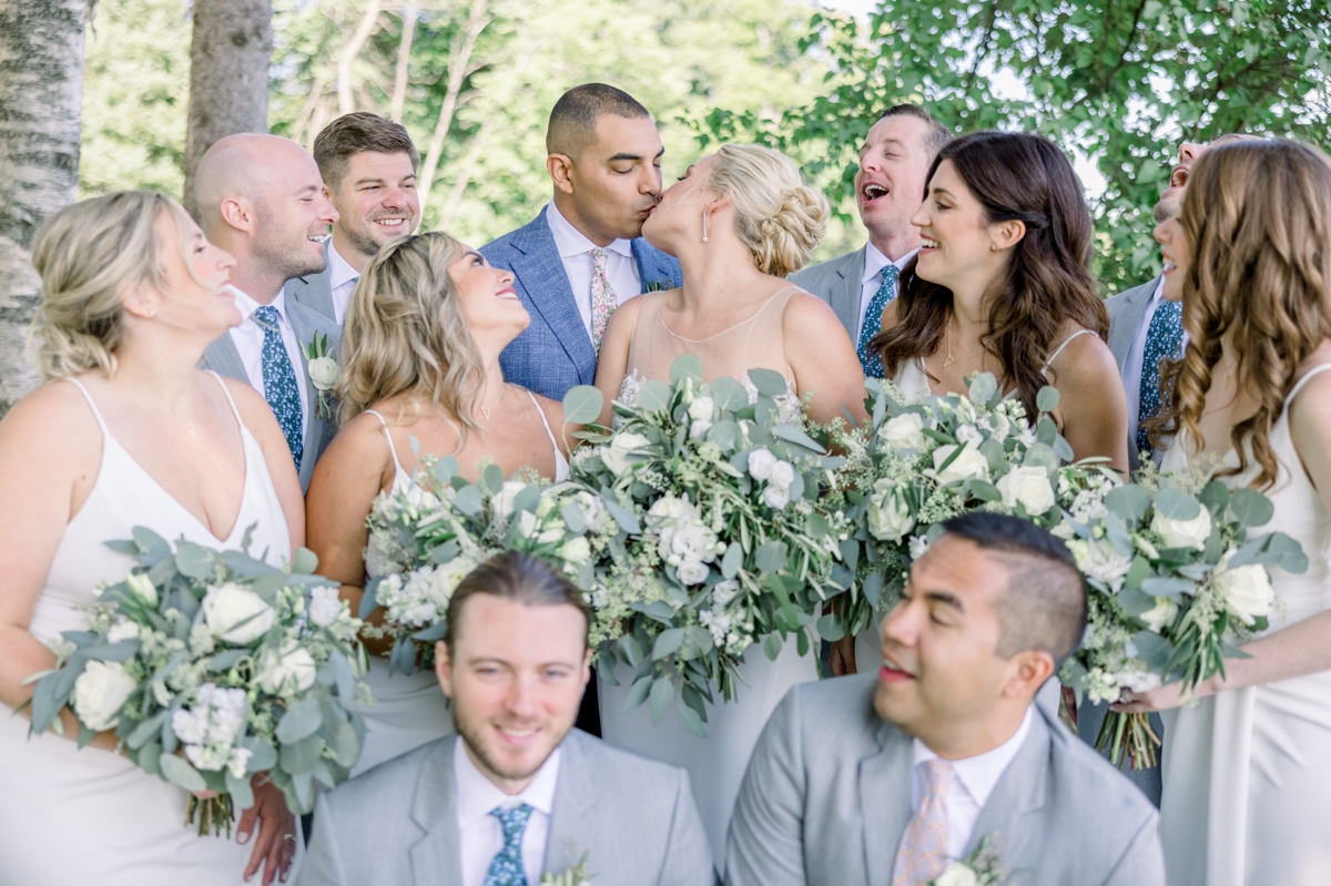 Caitlin and Andres kissing as their wedding party gathers around and smiles at them on their wedding day.