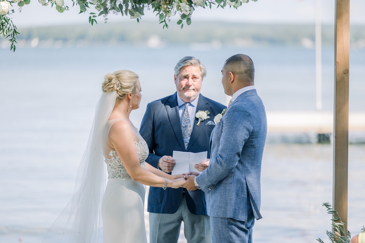 Caitlin and Andres holding hands as they both look at their officiant during their lakeside wedding ceremony.