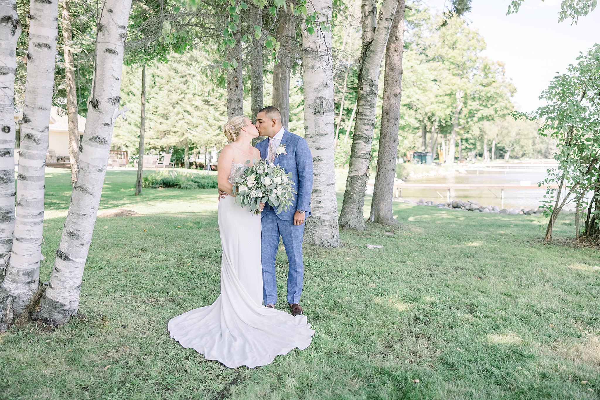 Andres kissing his bride under a grove of birch trees on their wedding day as they stand on the shore of Burt Lake in Northern Michigan